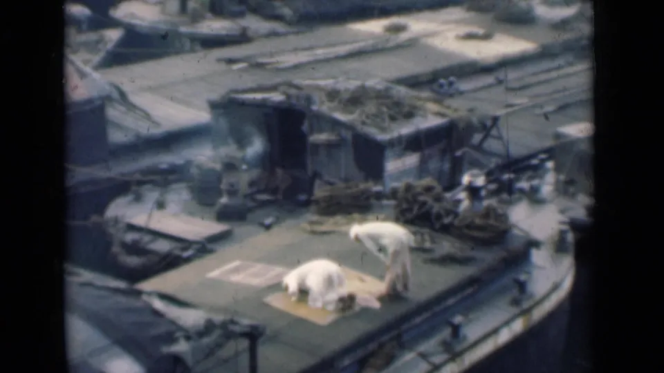 A high-angle 8mm frame from Calcutta shows two men in white on a deck, one kneeling low on a mat as the other bends nearby.
