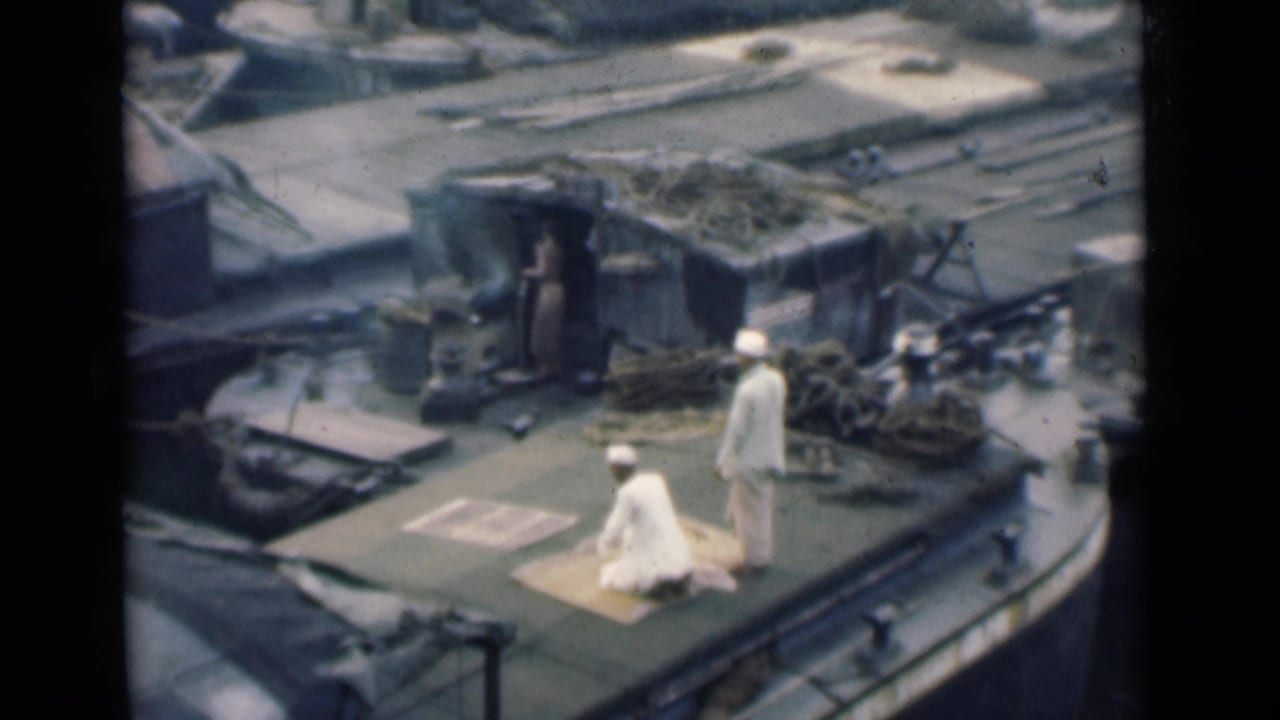 Two sailors in white uniforms stand near a dog on a ship deck with harbor water beyond.