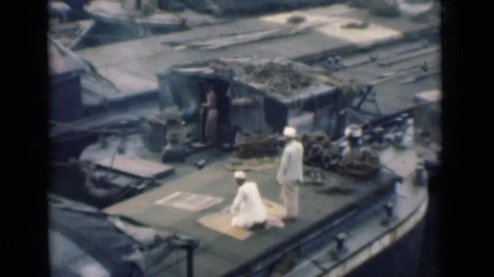 1940s home-movie view of two sailors on a cluttered ship deck near a white dog, with harbor water visible in the background.