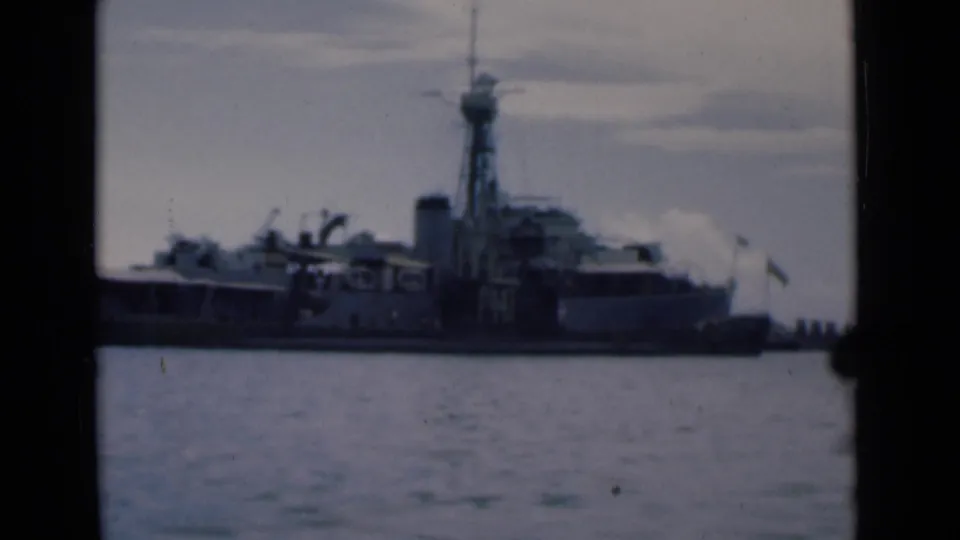 Faded 8mm frame showing a grey naval ship broadside at Colombo harbor with dock buildings and flags nearby.