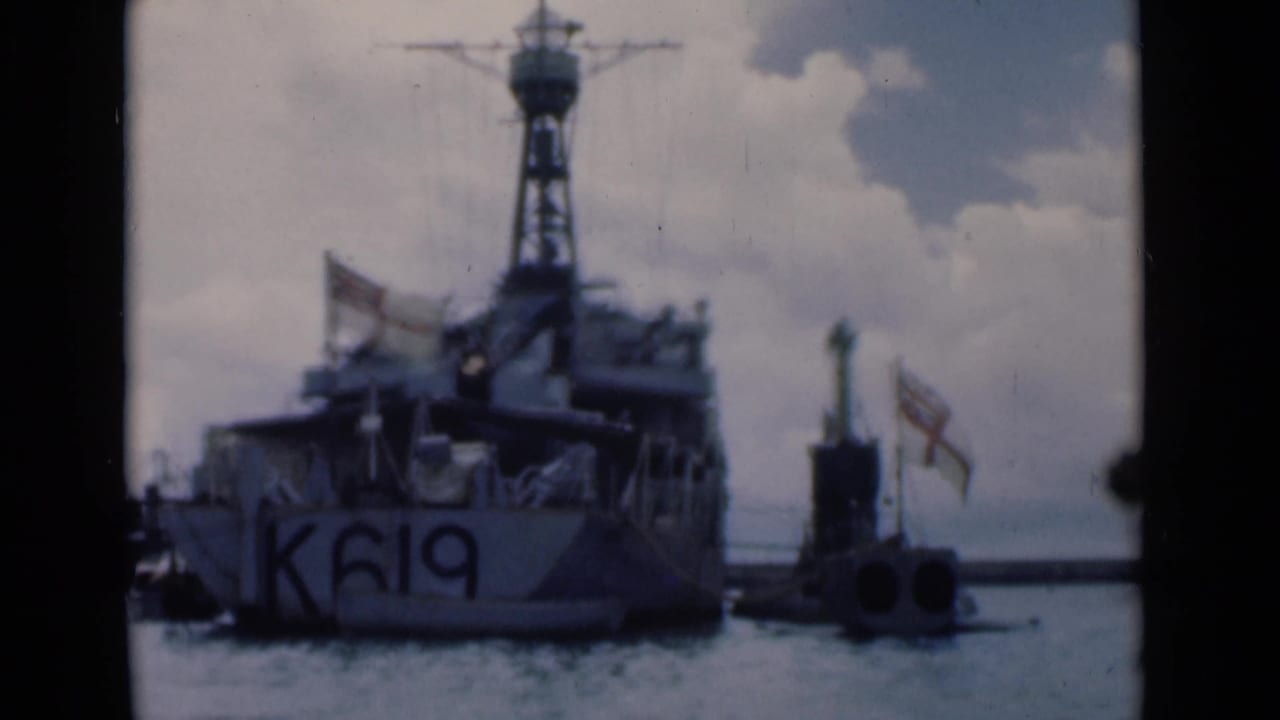 Stern of naval vessel K619 in harbor with flags and a small marker nearby.
