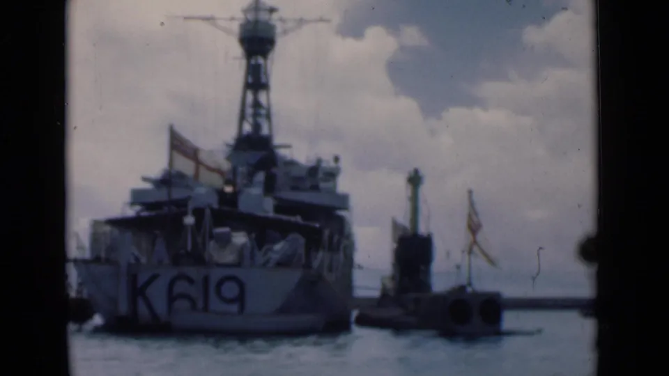 A soft-focus 8mm frame shows vessel K619 from the stern in Colombo harbor, with rigging, flags, water, and a low marker beside it.