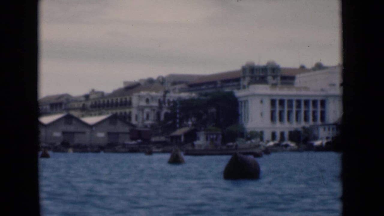 White waterfront building across blue harbor water with buoys and small boats.
