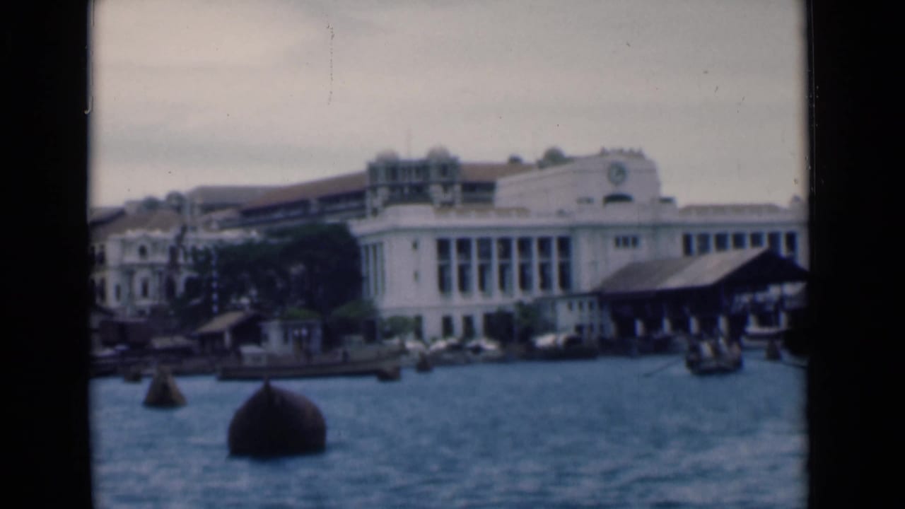 Harbor water and buoys in front of a large pale building and dock sheds.