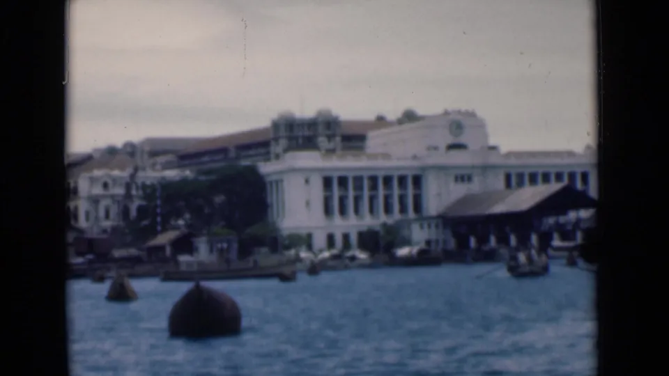 Boat-level view of Colombo waterfront architecture, with dark buoys, small craft, dock sheds, and a large pale building along the shore.