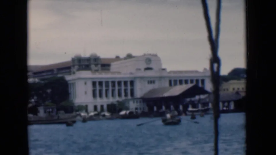 1946 Colombo waterfront seen from a moving boat, with a white building, dock shed, small boats, and rope or rigging crossing the frame.