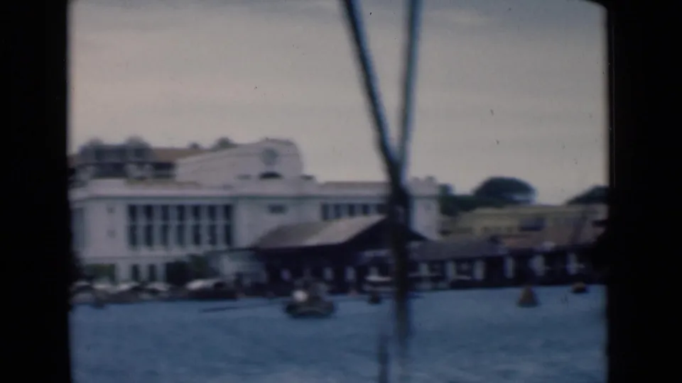 Soft 8mm frame of the Colombo waterfront, showing a large white building, dock shed, small boats, a buoy, and vertical rigging near the lens.