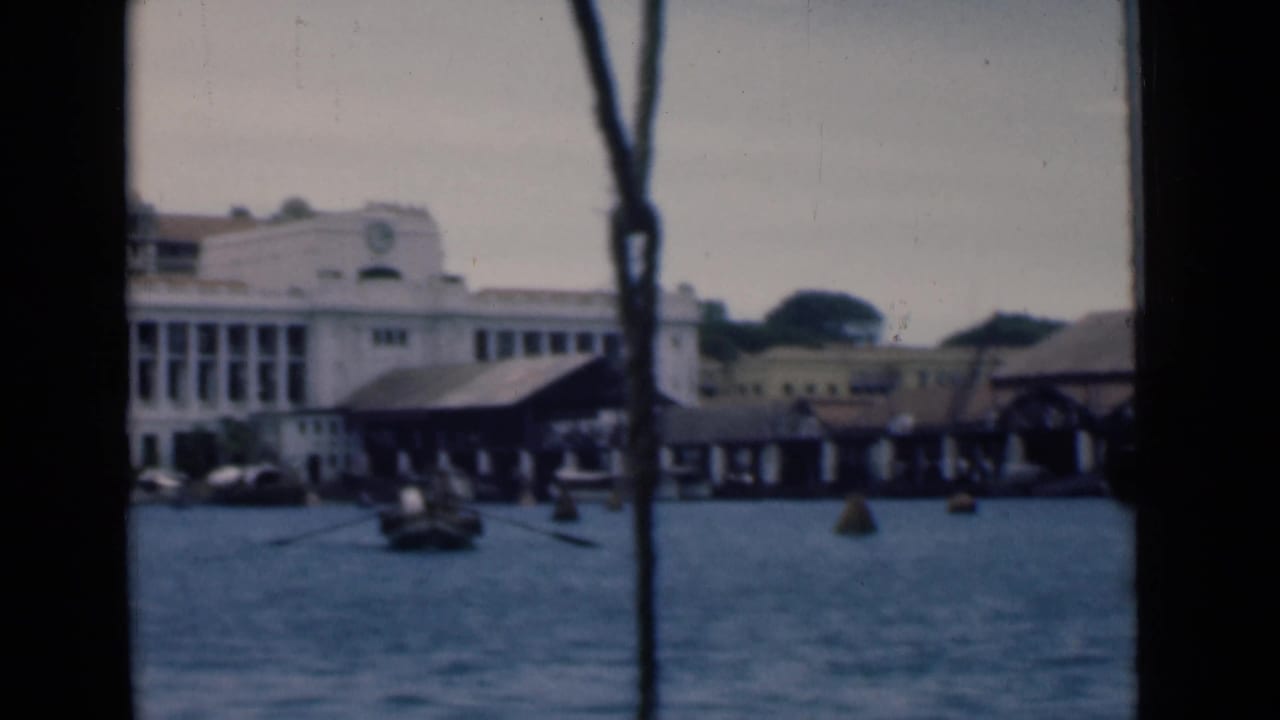 1946 Colombo harbor view with a white clock-tower building, covered docks, small boats, and dark boat framing in the foreground.