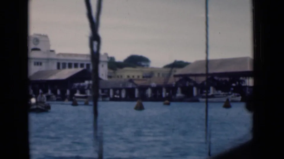 Moving 8mm view across Colombo waterfront docks, with a clock-tower building partly framed by dark vertical boat structures.