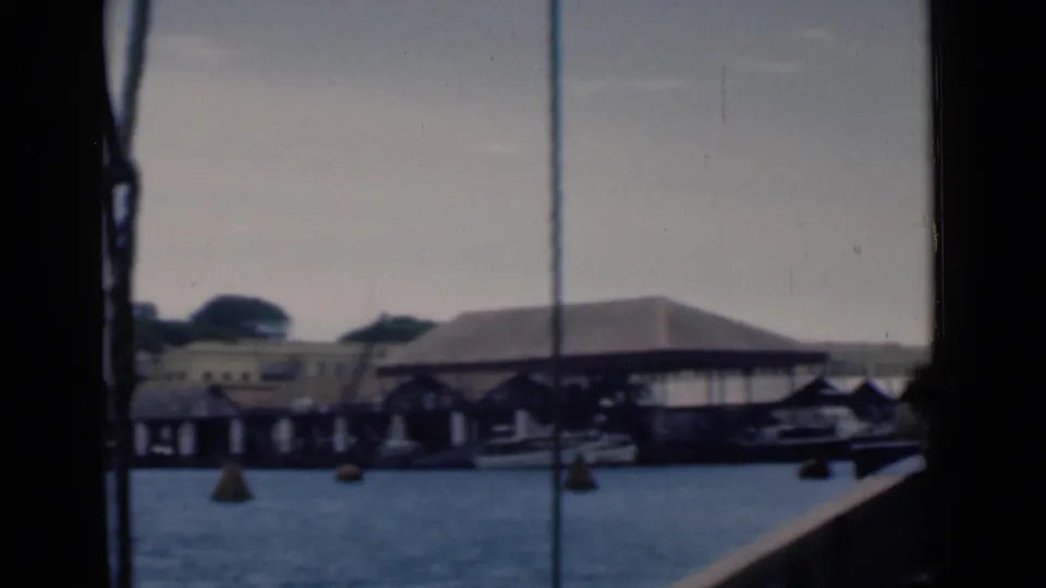 Colombo harbor frontage in 1946, showing covered pier structures, rooflines, buoys, small boats, and a partial boat rail in the foreground.