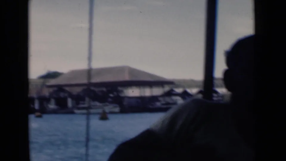 End frame of the 1946 Colombo boat view, with harbor water, covered dock buildings, buoys, and a dark passenger silhouette at the right edge.