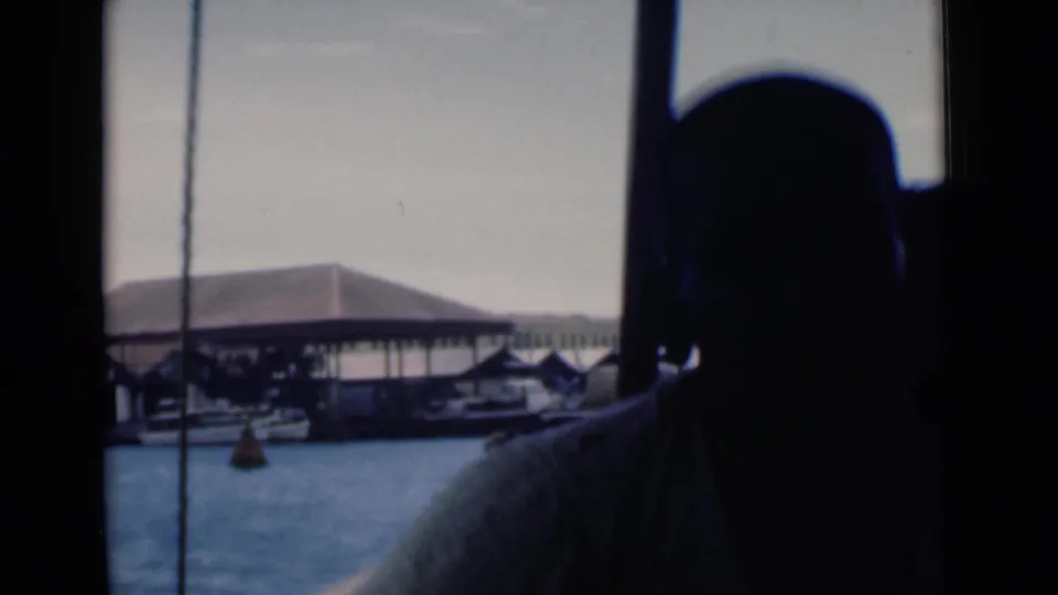 A silhouetted traveler on a boat looks out toward Colombo harbor structures and small boats in 1946 home-movie footage.