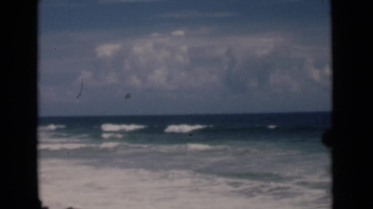 1946 8mm home-movie frame of surf rolling toward a Colombo, Ceylon shoreline under broken clouds, with dark gate edges visible.