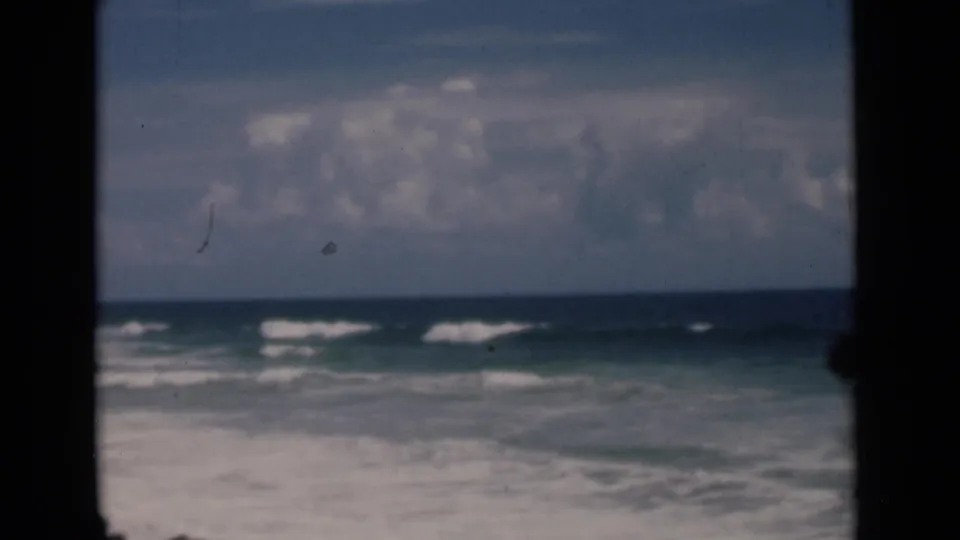 1946 8mm home-movie frame of surf rolling toward a Colombo, Ceylon shoreline under broken clouds, with dark gate edges visible.