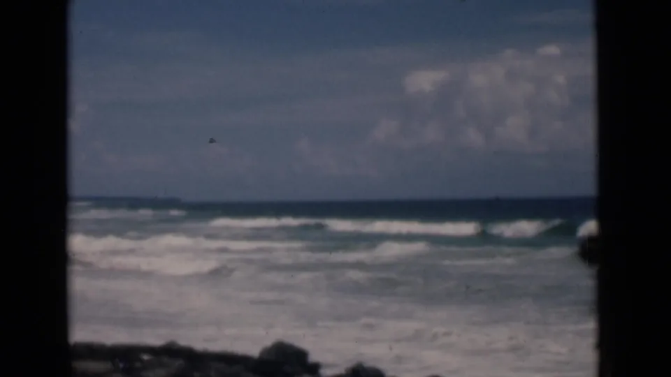 1946 8mm coastal frame from Colombo, Ceylon showing breaking surf, low horizon, clouds, and visible film dust near the center.