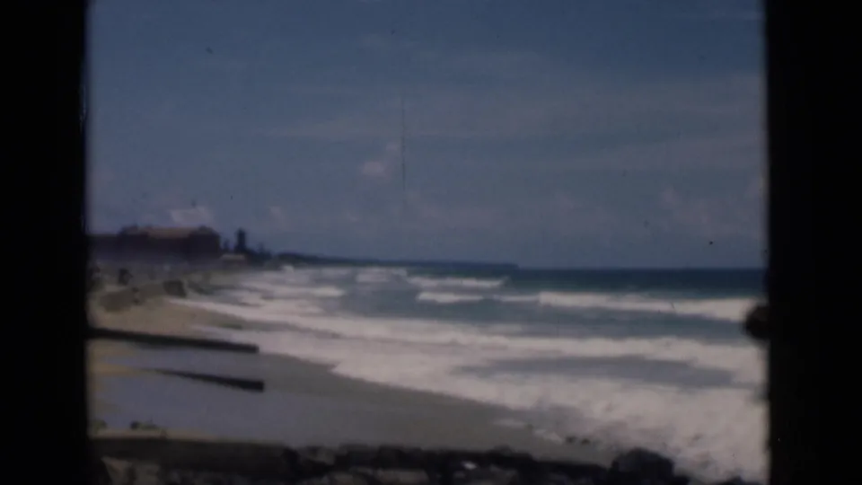 Colombo, Ceylon shoreline in 1946, with foamy waves, pale sand, distant coast, and dark framing from a shaded opening.