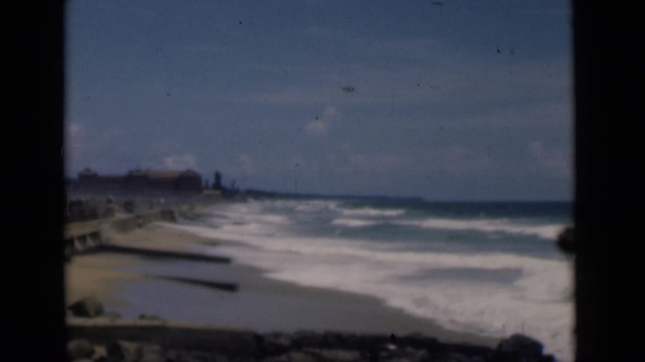 1946 Colombo, Ceylon coastal view with surf rolling toward a sandy beach beside a stone seawall and distant shoreline buildings.
