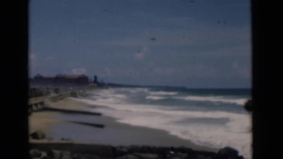 1946 Colombo, Ceylon coastal view with surf rolling toward a sandy beach beside a stone seawall and distant shoreline buildings.