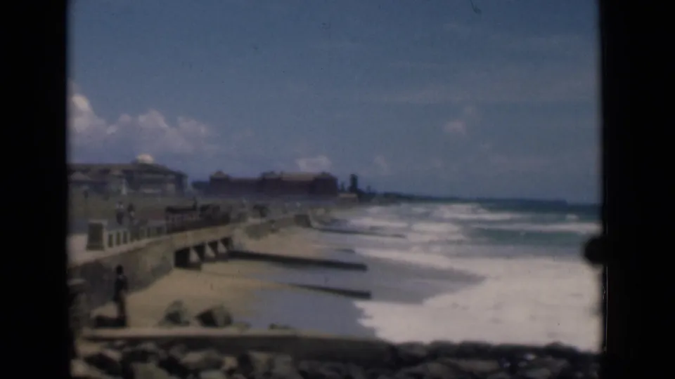 1946 home-movie view of a Colombo beach promenade with a seawall, rolling surf, rocks, and a few distant people.