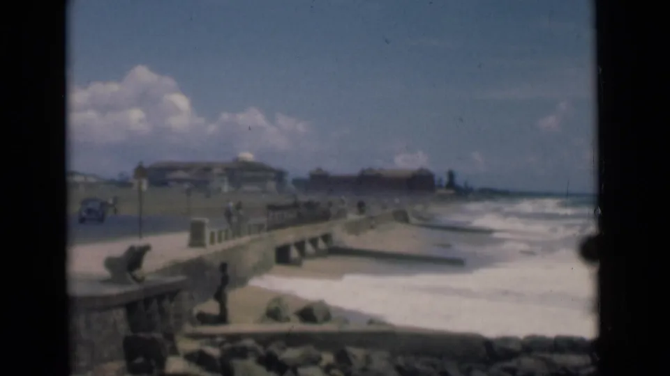1946 Colombo 8mm frame showing a seafront promenade, stone seawall, waves, an open road, and distant pedestrians near low buildings.