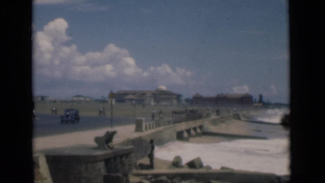 Seafront road beside a stone promenade with surf, pedestrians, a car, and distant buildings.