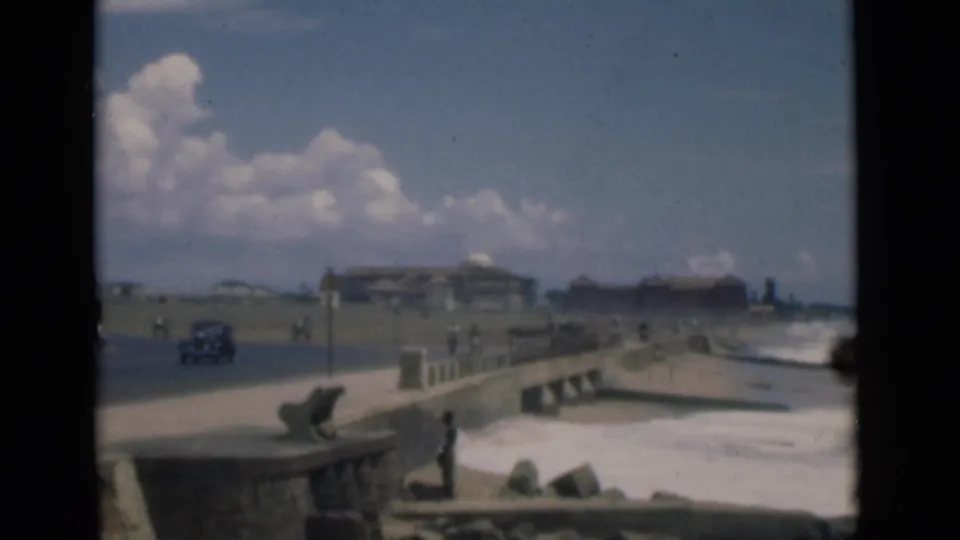 1946 Colombo coastal street view with a stone promenade, ocean waves, pedestrians, a vintage car, and distant seafront buildings.