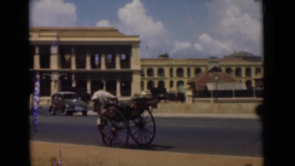 1946 Colombo street frame showing a man with a hand-pulled rickshaw, classic cars, and colonial-era buildings with columns and arched windows.