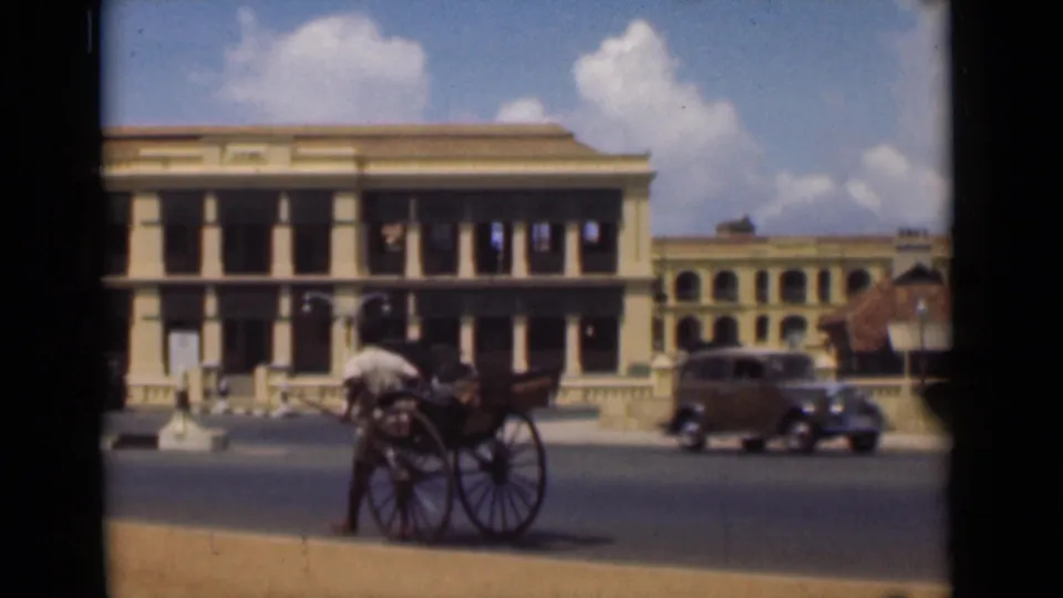 1946 Colombo street scene with a rickshaw puller, hand-pulled rickshaw, passing vintage car, and colonial building under a bright sky.