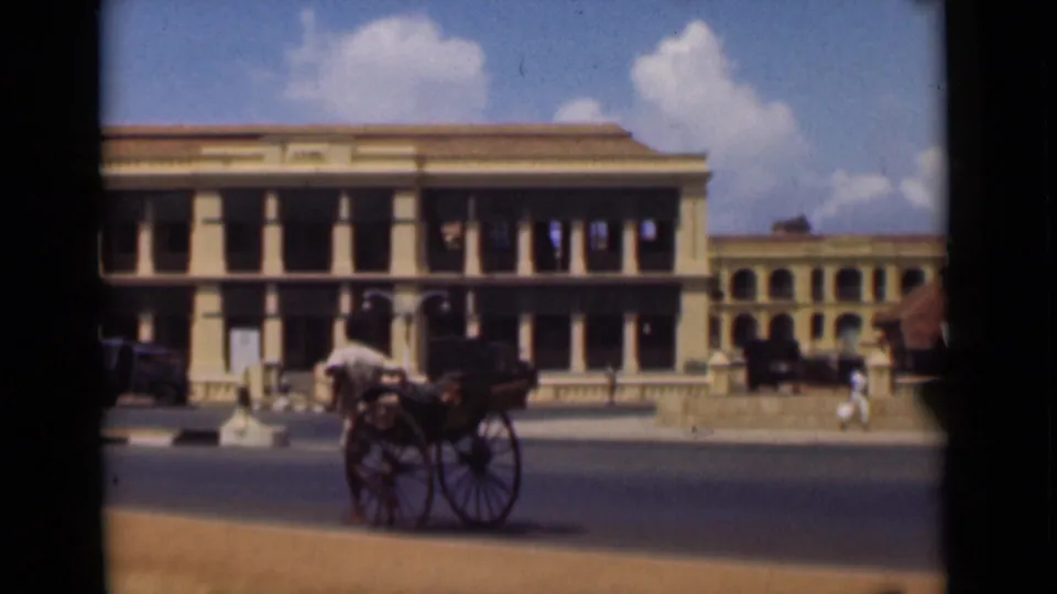 In 1946 Colombo, a man pulls a two-wheeled cart along a road in front of columned colonial-era buildings.
