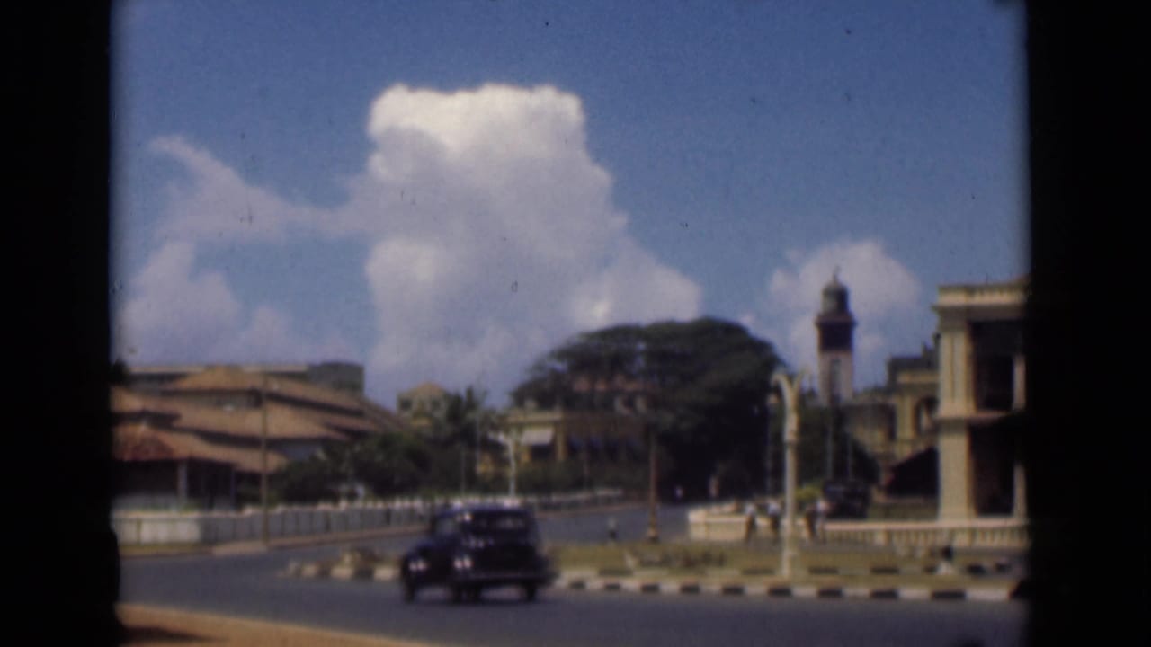 Classic car driving near a clock tower and columned buildings under large clouds.