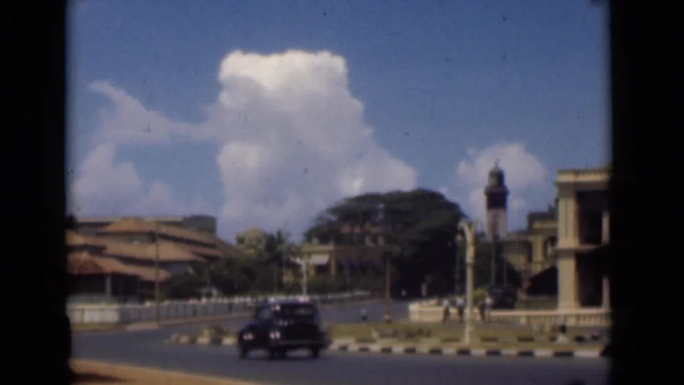 A classic automobile passes through a Colombo street scene with a clock tower, railings, trees, and colonial-era buildings.