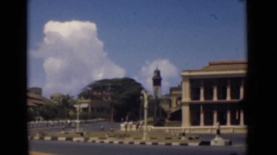 The final frame holds on the Colombo streetscape, with a clock tower, railings, trees, road markings, and a columned building.
