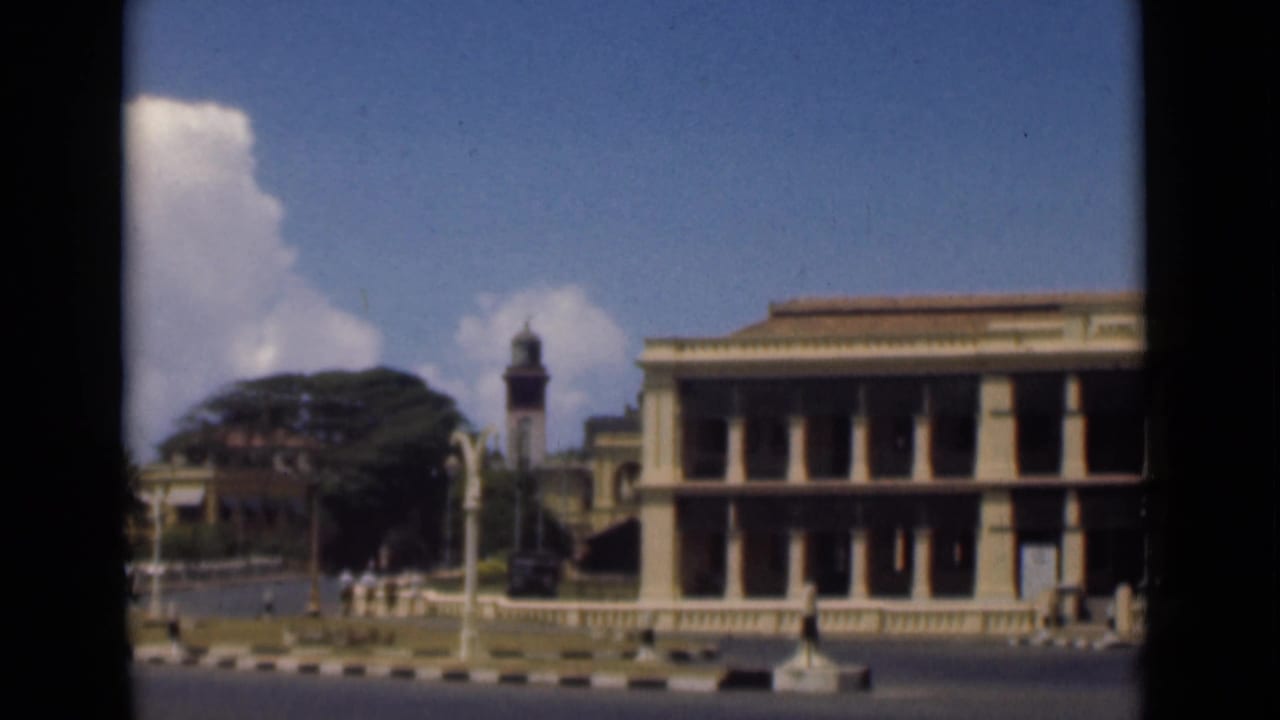 Street scene with a colonnaded building, clock tower, trees, and small pedestrians.