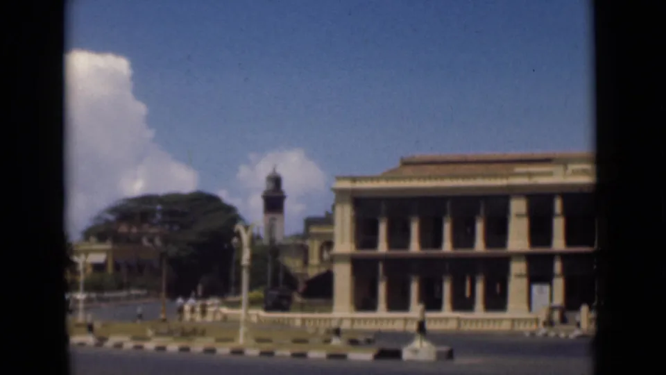 1946 Colombo street scene filmed from a moving vehicle, showing a colonnaded facade, clock tower, trees, and distant pedestrians.