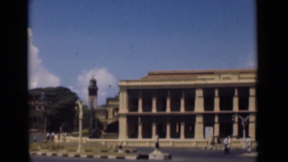 1946 Colombo view of a large colonnaded building along a quiet street, with pedestrians, a clock tower, streetlamps, and clouds.