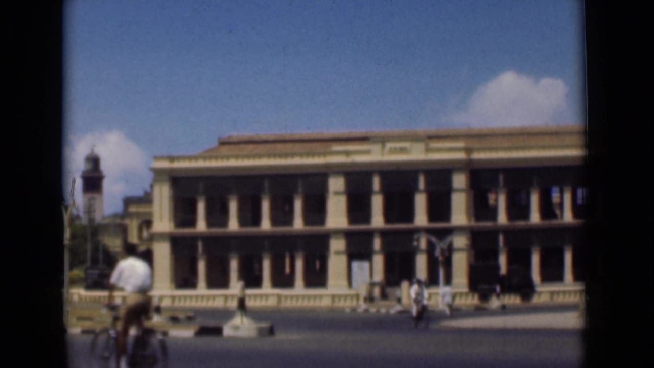 Cyclist passes in front of a long columned building on a broad city street.