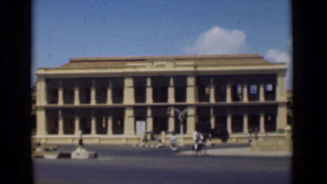 Pedestrians and bicycles appear near a long building with columns and a shaded upper level.