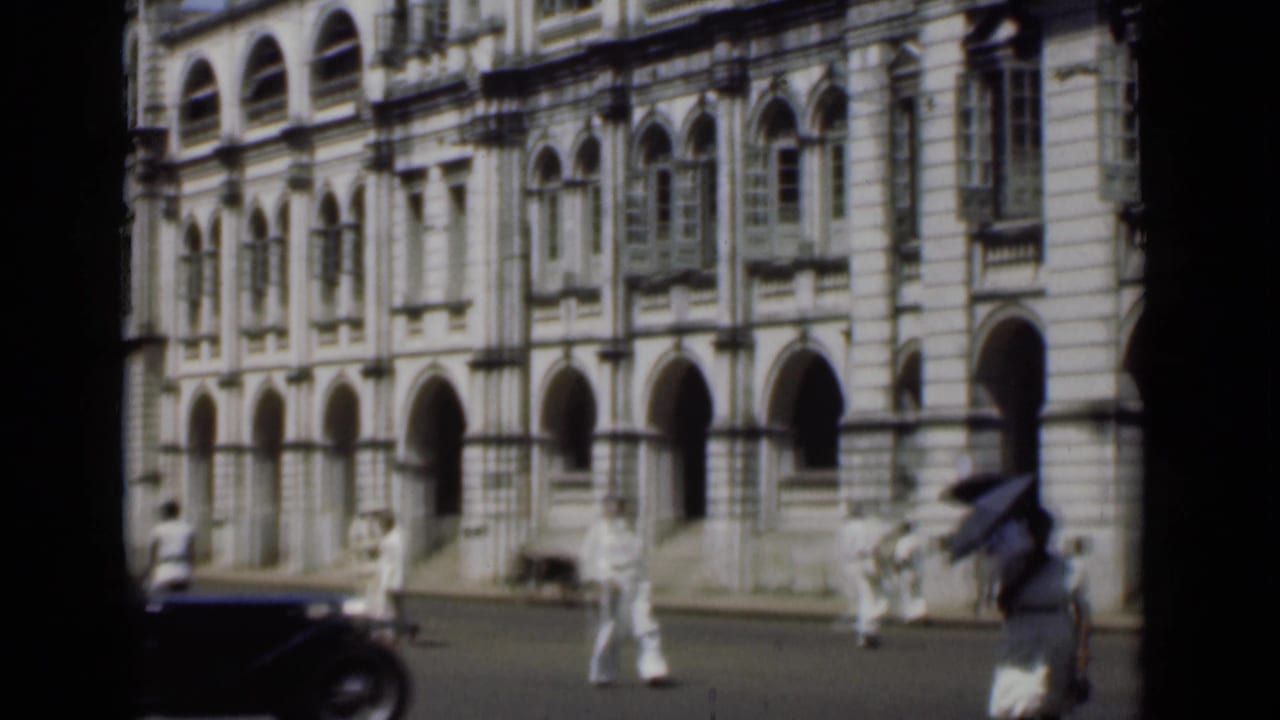 Pedestrians walk past a large arched building while a dark car enters the street foreground.