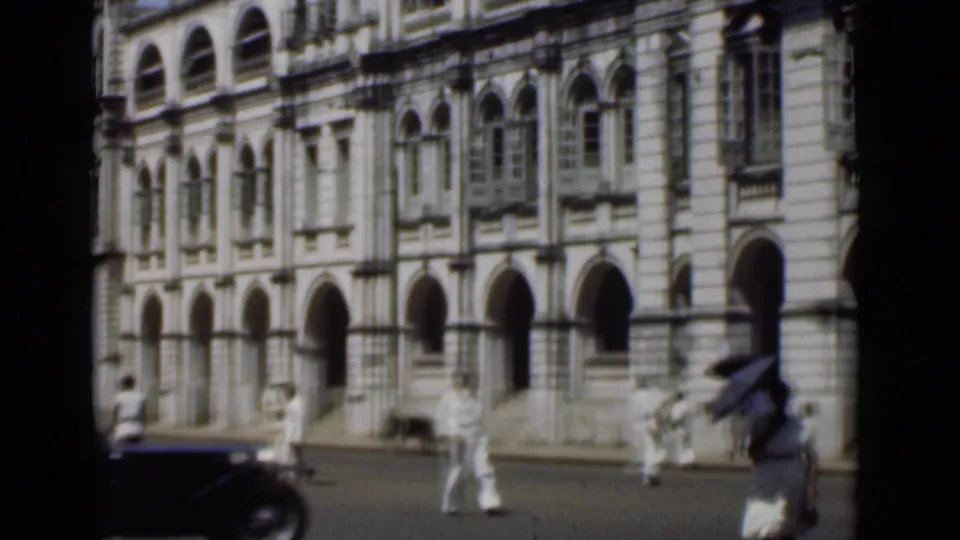 1946 Colombo street view with pedestrians near a grand arched facade and a dark vintage car at the edge of frame.