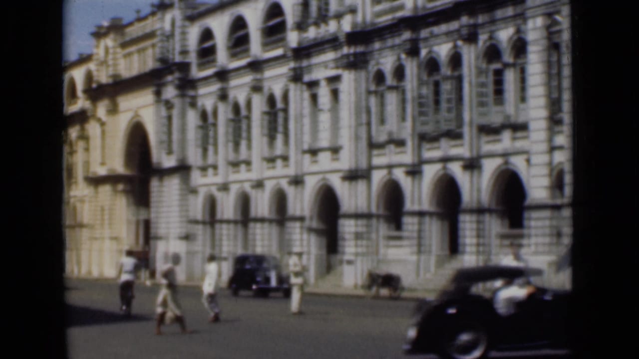 People cross an open street in front of an ornate building as a black car passes nearby.
