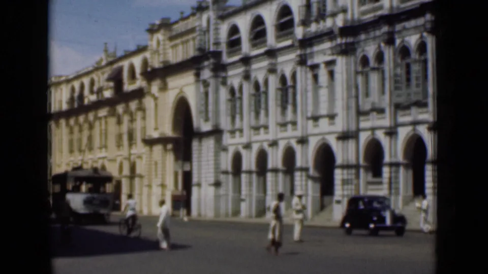 1946 Colombo view with pedestrians, a vehicle at the left edge, and a dark car beside a large colonial-style facade.
