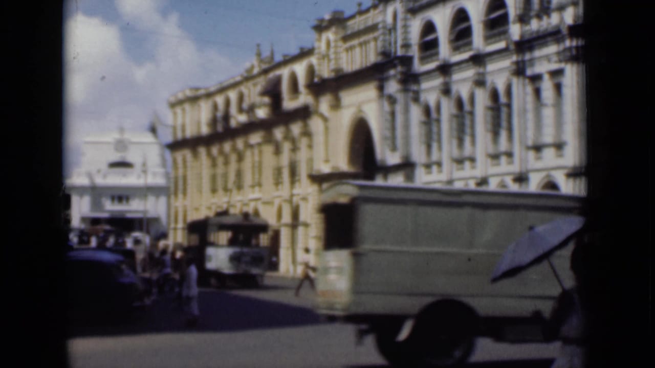 Vintage street with delivery truck, pedestrians, cars, arched buildings, and a white building beyond.