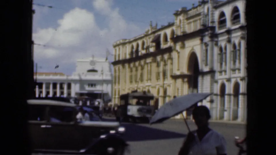A sedan passes through a 1946 Colombo street as pedestrians and an umbrella-holder move near colonial-era commercial buildings.