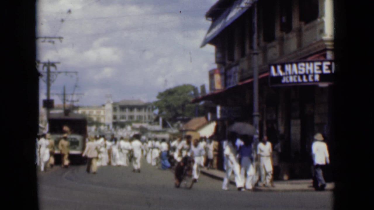 Crowded street with pedestrians, shopfront sign, utility poles, and a vehicle in the distance.