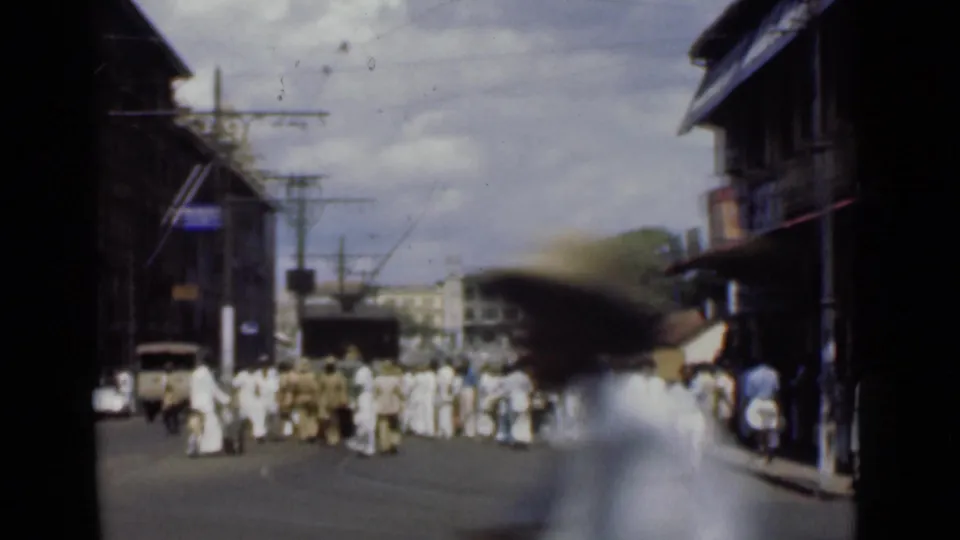 1946 Colombo, Ceylon street view with a blurred passerby close to camera and pedestrians gathered across the road near overhead utility lines.