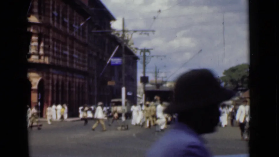 1946 Colombo, Ceylon street scene showing pedestrians crossing near colonial-style facades, overhead wires, and a silhouetted passerby in the foreground.