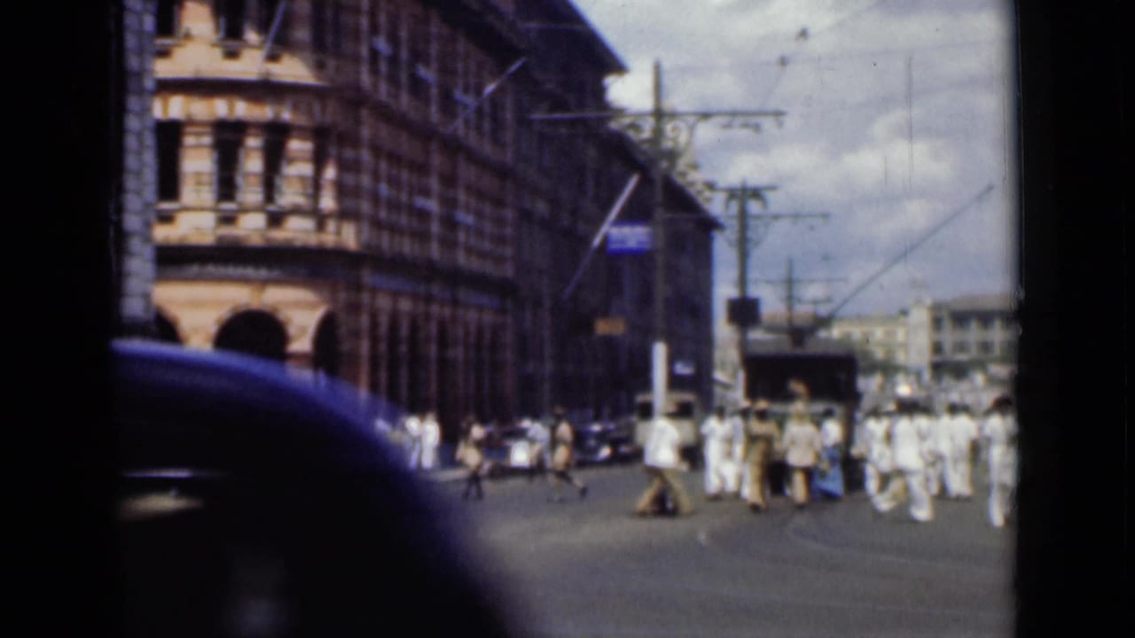 Busy city street with pedestrians, tall masonry buildings, overhead wires, and a dark car foreground.