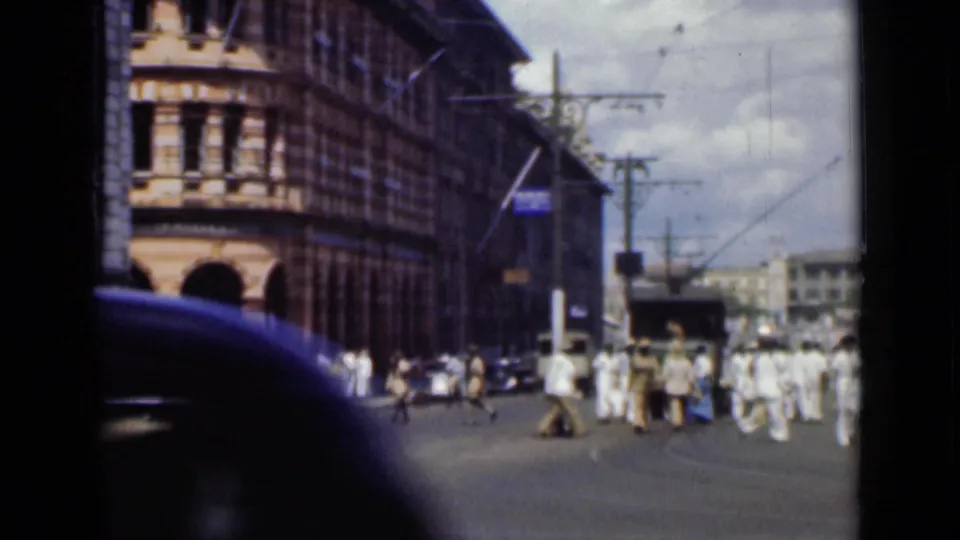 1946 Colombo street scene filmed from a vehicle, with pedestrians crossing near tall colonial-era buildings and a dark car edge in front.