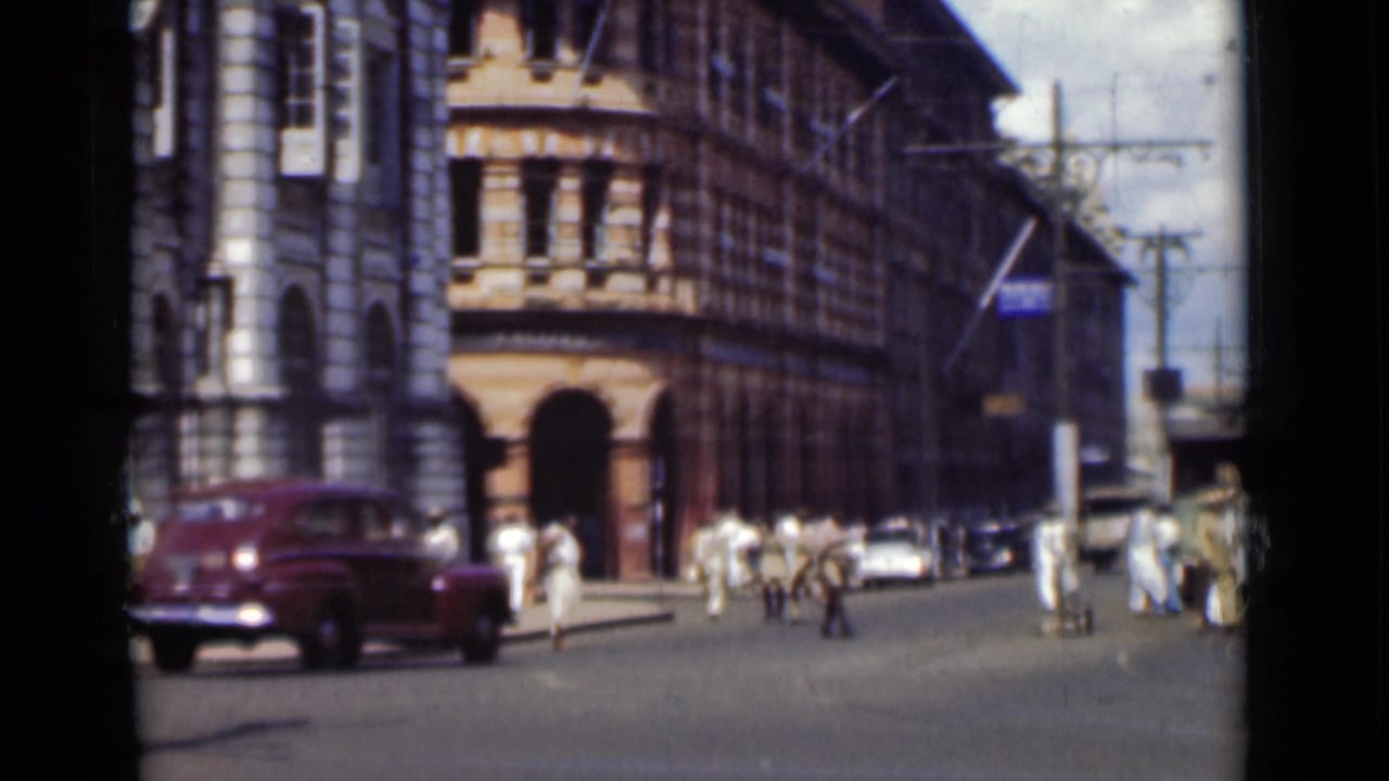 A maroon mid-century car drives through a Colombo intersection as pedestrians move near ornate urban buildings.
