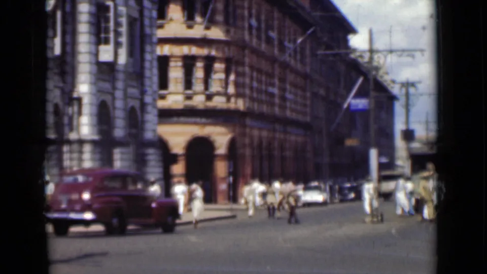 A maroon mid-century car drives through a Colombo intersection as pedestrians move near ornate urban buildings.